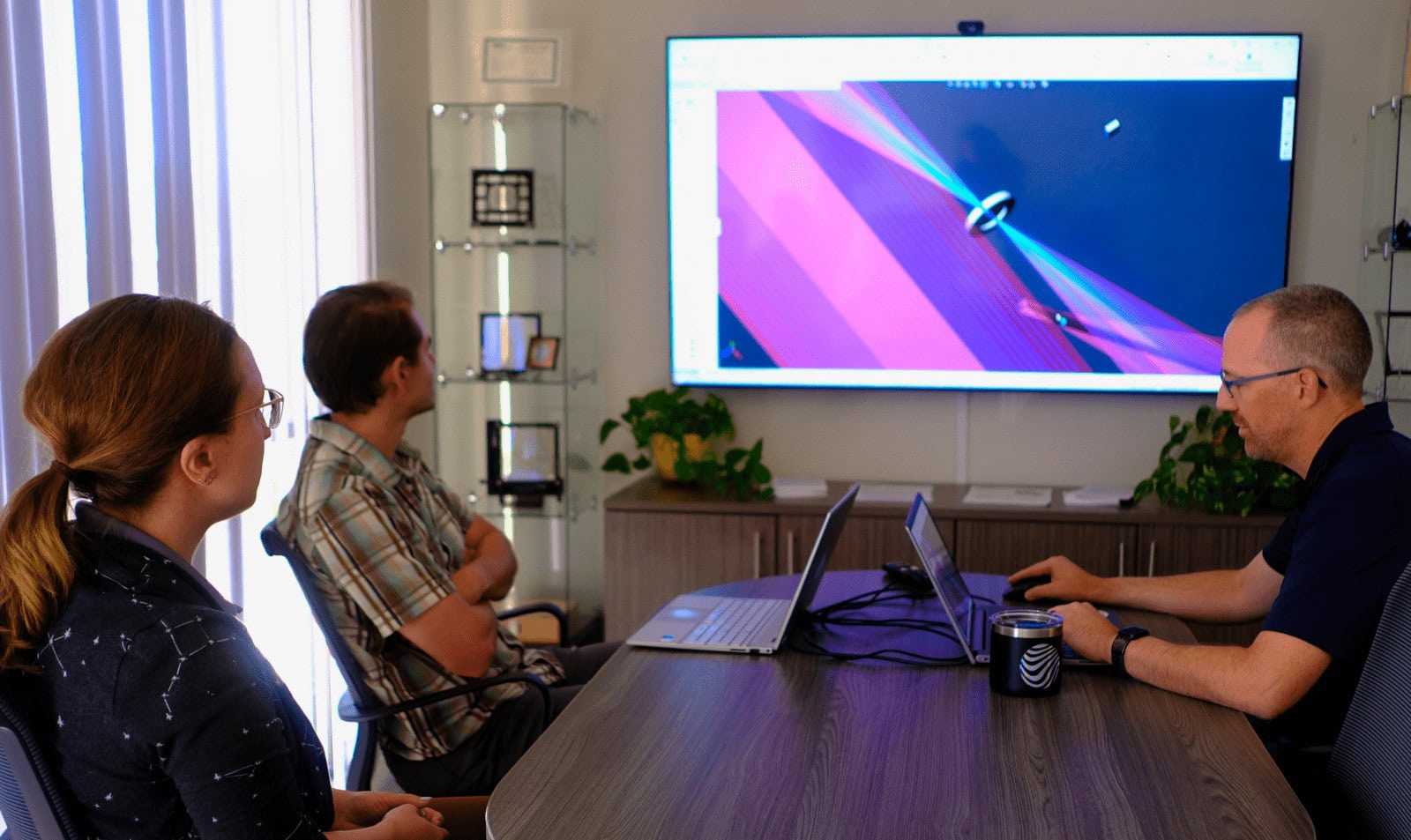 Three engineers sitting at a conference table analyze a ray diagram of an optical system on a large TV screen